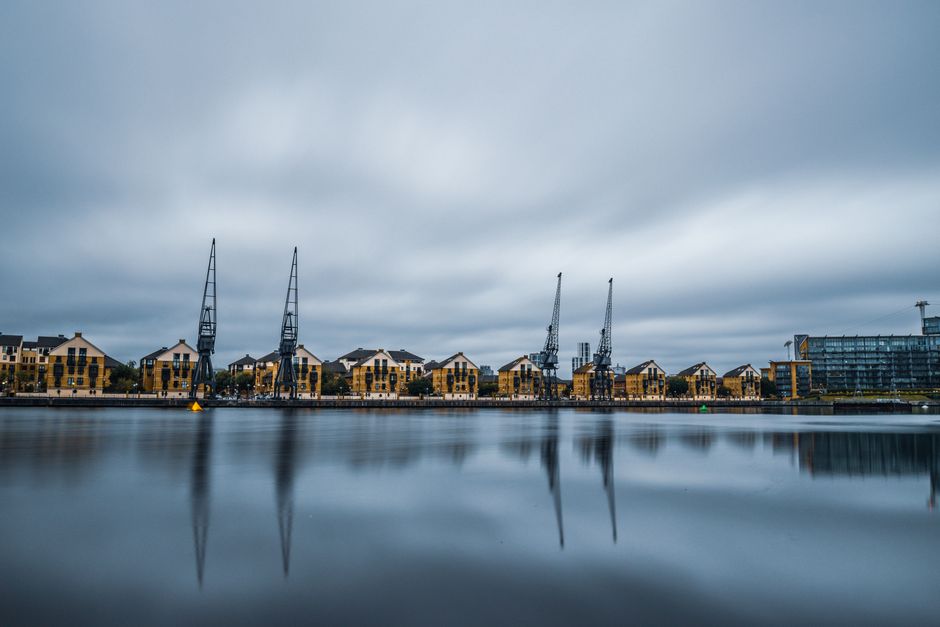 View of a dockside area in Docklands showing a row of modern yellow and beige residential buildings along the waterfront. Four large industrial cranes with tall, dark metal structures are positioned behind the buildings, suggesting a working port or shipyard. The scene is captured under a cloudy, grey sky, with diffused lighting creating a calm atmosphere. The calm water in the foreground reflects the buildings and cranes, enhancing the visual symmetry. The surroundings include paved walkways and some greenery near the buildings. This image relates to the broader context of property and home relocation services offered by Docklands Removals, illustrating the area where furniture and belongings may be transported during a move, with the port infrastructure hinting at the logistics involved in moving household items across the region.