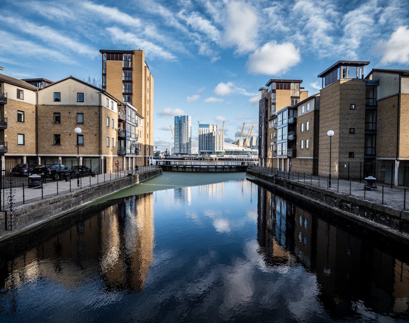 A wide-angle view of a waterfront in the Docklands area showing modern high-rise buildings with glass facades reflecting sunlight, including a prominent curved residential tower. In the foreground, there is a small marina with wooden floating docks, some of which are equipped with red and black protective bumpers. Several people can be seen walking along the promenade, which is lined with trees and benches, suggesting a public space used for leisure and pedestrian access. The sky overhead is partly cloudy with patches of blue, and the water surface shows gentle ripples. The image captures the urban environment during daylight hours, illustrating the mix of commercial, residential, and recreational spaces typical of a city’s waterfront. Docklands Removals may use this setting to transport furniture and household belongings through similar modern, urban landscapes during home relocation or packing and moving processes.