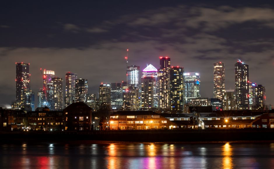 Night-time view of the Canary Wharf skyline in London with illuminated modern skyscrapers featuring glass facades, some topped with red aviation warning lights. In the foreground, there is a row of low-rise residential buildings with lit windows, situated along the waterfront reflecting the city's lights on the dark water surface. The cloudy sky above adds to the urban night scene. This image visually depicts the busy high-rise environment typical of central London’s docklands area, relevant for house removals and furniture transport services offered by Docklands Removals for E14 flats and the surrounding area.