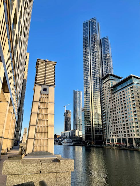 A view of a cityscape along a waterway in Canary Wharf, featuring modern high-rise office buildings with glass facades and steel structures, under a clear blue sky. In the foreground, there is a stone platform with a tall, rectangular metal vent or chimney, positioned on a paved area close to the water. The scene depicts The docks and commercial district with skyscrapers and construction cranes visible in the distance, illustrating the ongoing development and urban environment associated with house removals and furniture transport in E14 flats. Docklands Removals can assist with the logistics of packing, loading, and relocating furniture and boxes within the busy Canary Wharf area, as shown in the context of home relocation and moving services.