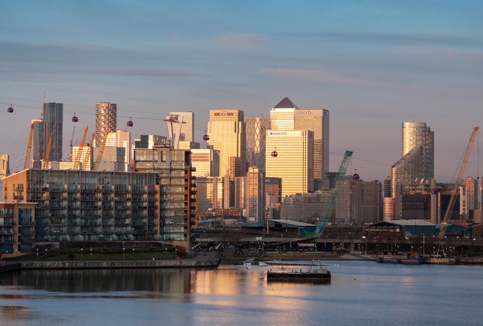Night-time view of the Canary Wharf skyline in London with illuminated modern skyscrapers featuring glass facades, some topped with red aviation warning lights. In the foreground, there is a row of low-rise residential buildings with lit windows, situated along the waterfront reflecting the city's lights on the dark water surface. The cloudy sky above adds to the urban night scene. This image visually depicts the busy high-rise environment typical of central London’s docklands area, relevant for house removals and furniture transport services offered by Docklands Removals for E14 flats and the surrounding area.