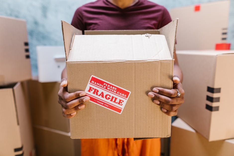 A person holding a medium-sized cardboard box labeled 'fragile' with red and white stickers that read 'Please handle with care' and 'Thank you,' inside a room filled with packing supplies. The individual is gripping the box from both sides, standing amidst other closed cardboard boxes stacked and arranged on the floor and shelves. The background shows more boxes, some with black tape, and part of a door or doorway, indicating a home environment prepared for a move. This image reflects careful packing and handling processes typical of home relocation services, such as those offered by Docklands Removals, especially for narrow access homes on the Isle of Dogs, where precise logistics and careful furniture transport are essential.