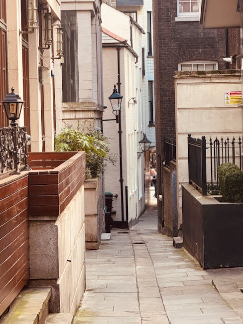 A narrow residential alleyway with a paved stone pathway leading between closely spaced buildings on either side. On the left, a wooden planter box with a small green shrub is positioned atop a stone wall beneath an ornate black metal lantern attached to a white building. Further along, a second black lantern is mounted on the same white façade. On the right, a tall black metal fence with pointed finials encloses a small garden area with a well-maintained shrub, and a beige stone wall supports a similar lantern. The alleyway appears to be part of a home relocation process, possibly accessed by a moving team from Docklands Removals, with the environment prepared for furniture and box transportation. The lighting suggests daylight, and the scene captures the tight spatial constraints typical of narrow access homes on the Isle of Dogs, making it representative of challenging urban moving logistics.