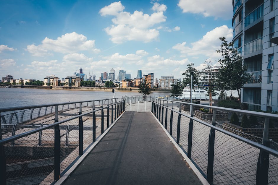 A wide, metal-railed pedestrian walkway extends along the edge of a river in Docklands, with a view of the city skyline featuring modern skyscrapers in the distance under a partly cloudy blue sky. To the right, a contemporary building with large windows and balconies is visible, along with small trees planted along the walkway. The walkway appears to be part of a home relocation or moving route, possibly used for transporting furniture or packing materials to a nearby relocations vehicle or storage area, reflecting the logistics involved in house removals in Docklands. The surface of the path is paved, and there are no people present in this scene, emphasizing the calm, organized environment typical of professional removals logistics managed by companies such as Docklands Removals.