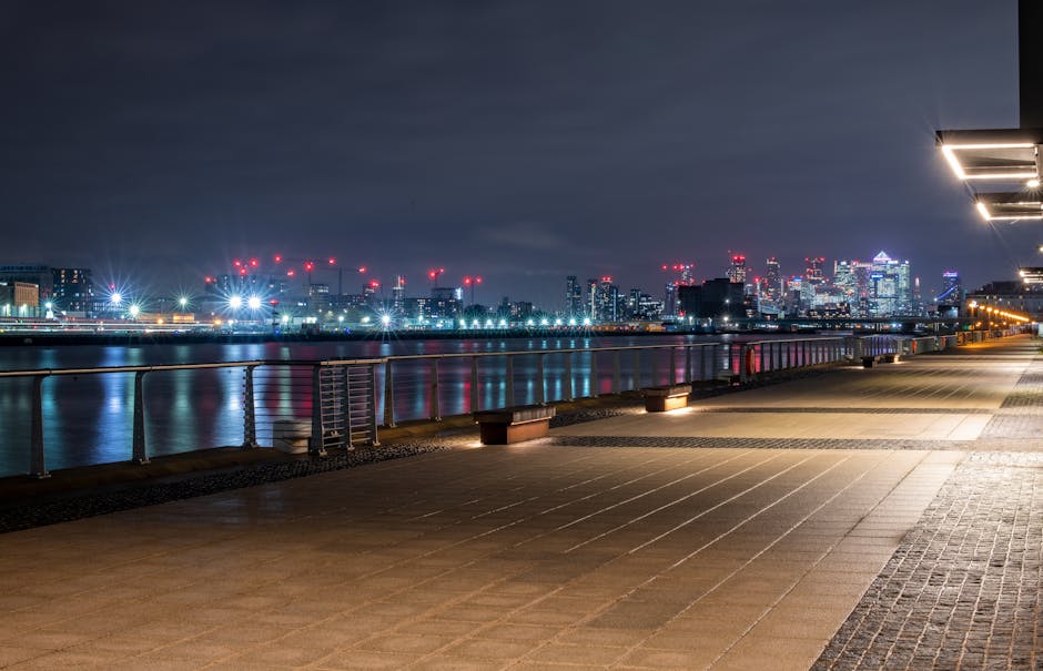 A wide-angle view of a riverside scene in Docklands featuring a row of modern, yellow residential buildings with dark roofs situated along the water's edge. In front of the buildings, several large cargo cranes are positioned on the quay, indicative of a working dock or port area. The foreground shows a calm, reflective body of water capturing the mirrored image of the buildings and cranes, with slight ripples distorting the reflection. The sky above is overcast with grey clouds, creating a subdued lighting atmosphere typical for a cloudy day. This setting illustrates an urban waterfront environment that could be associated with home relocation or furniture transport activities, often coordinated by removals companies like Docklands Removals, especially during loading and unloading phases of house moves or moving logistics.
