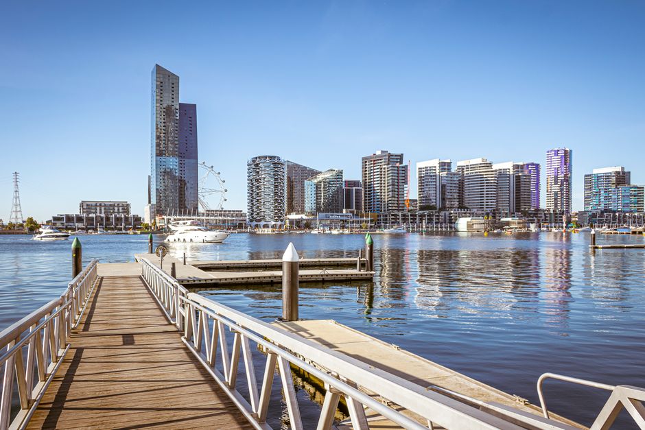 A photograph of a waterfront pier extending from the foreground towards a modern city skyline across the water, featuring high-rise buildings with glass facades, a Ferris wheel, and various commercial