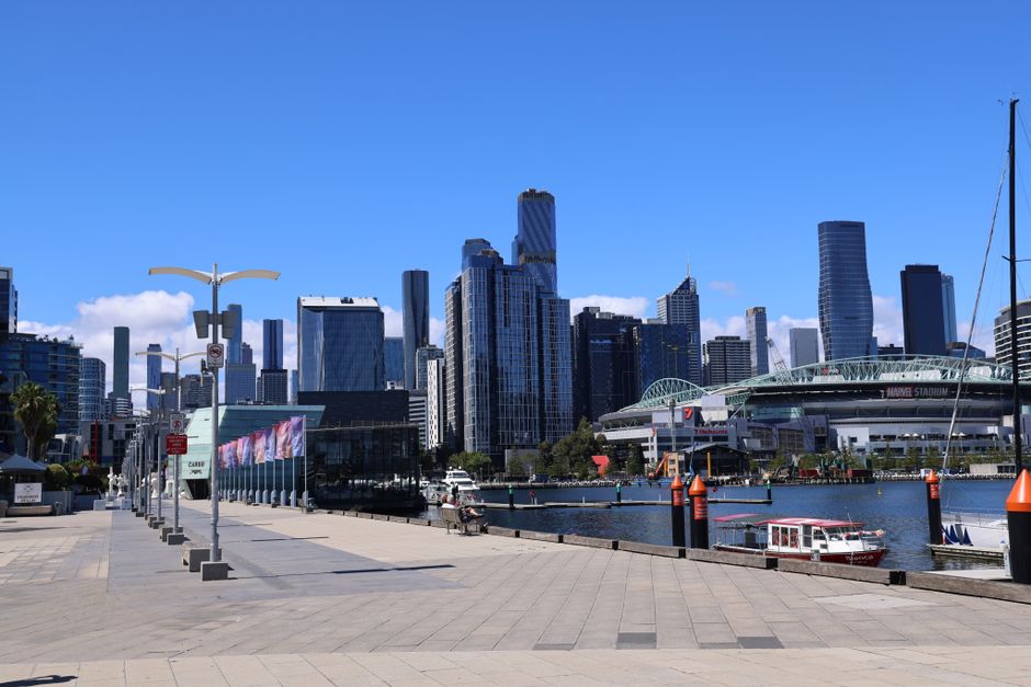 A photograph of a waterfront pier extending from the foreground towards a modern city skyline across the water, featuring high-rise buildings with glass facades, a Ferris wheel, and various commercial