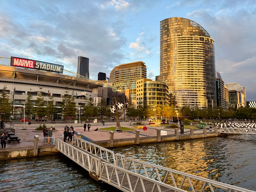 A panoramic view of Docklands with modern high-rise buildings along the waterfront, including several glass-fronted skyscrapers and a distinctive twisted tower, reflected clearly in the calm water bel